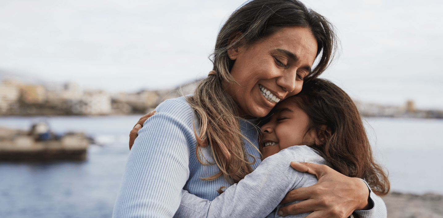 mother and daughter smiling and hugging outside by the water